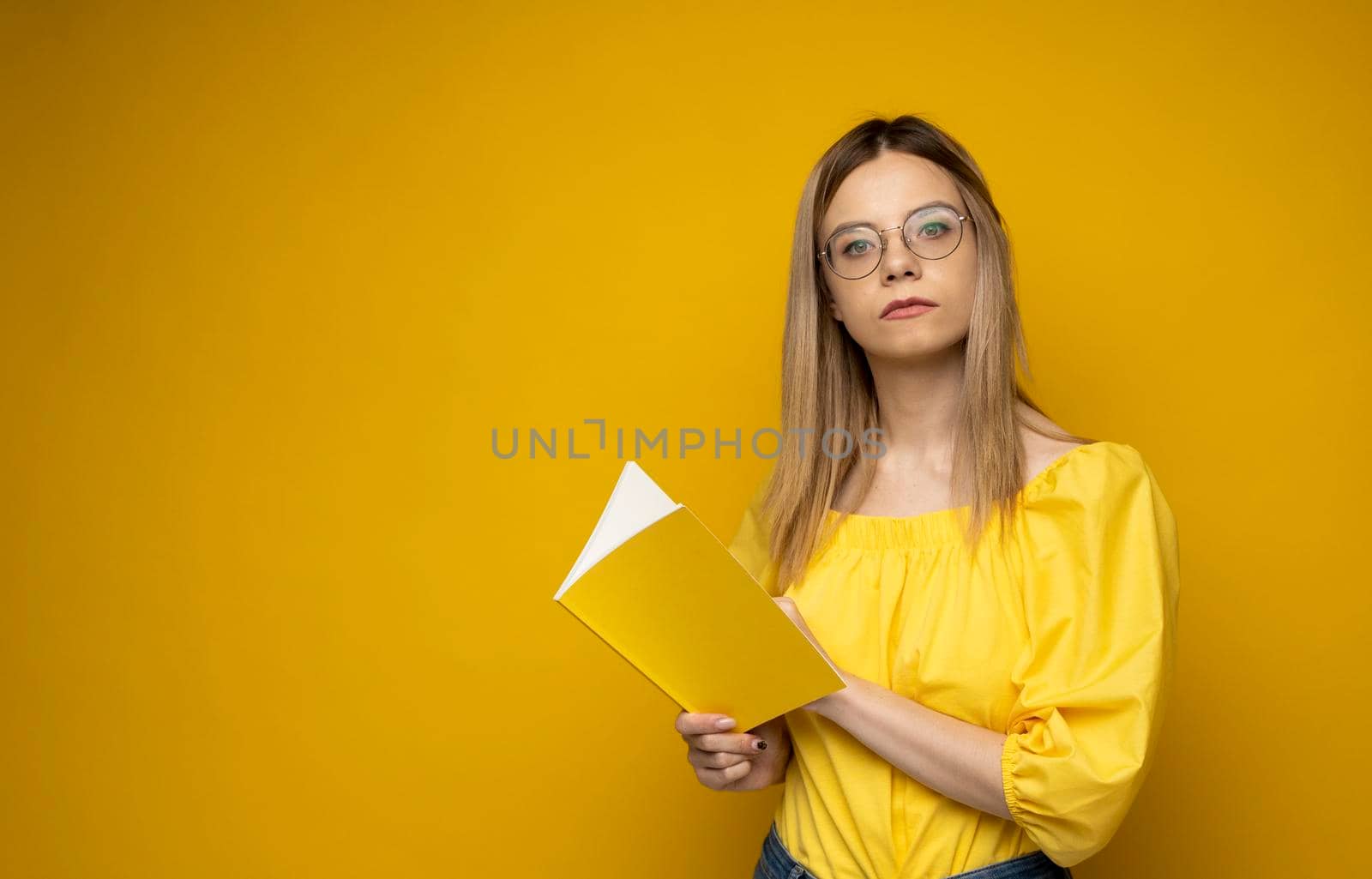 Beautiful smart young girl holding and reading book isolated on the yellow background. Portrait of attractive woman in a yellow blouse and wearing glasses reading book. Education, studying, knowledge. by vovsht