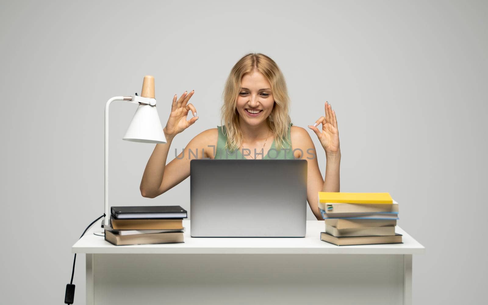 Student girl showing ok gesture with both hands, happy with completed work, smiling and looking at the camera while working on laptop computer sitting in front of big piles of books