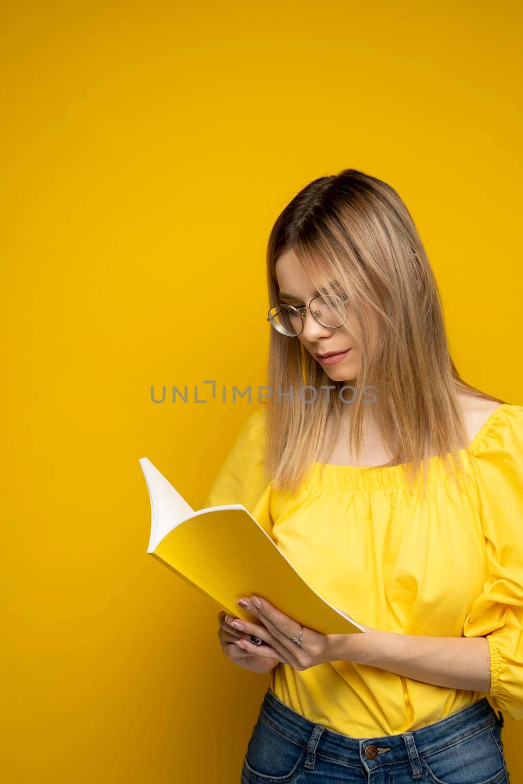 Beautiful smart young girl holding and reading book isolated on the yellow background. Portrait of attractive woman in a yellow blouse and wearing glasses reading book. Education, studying, knowledge. by vovsht
