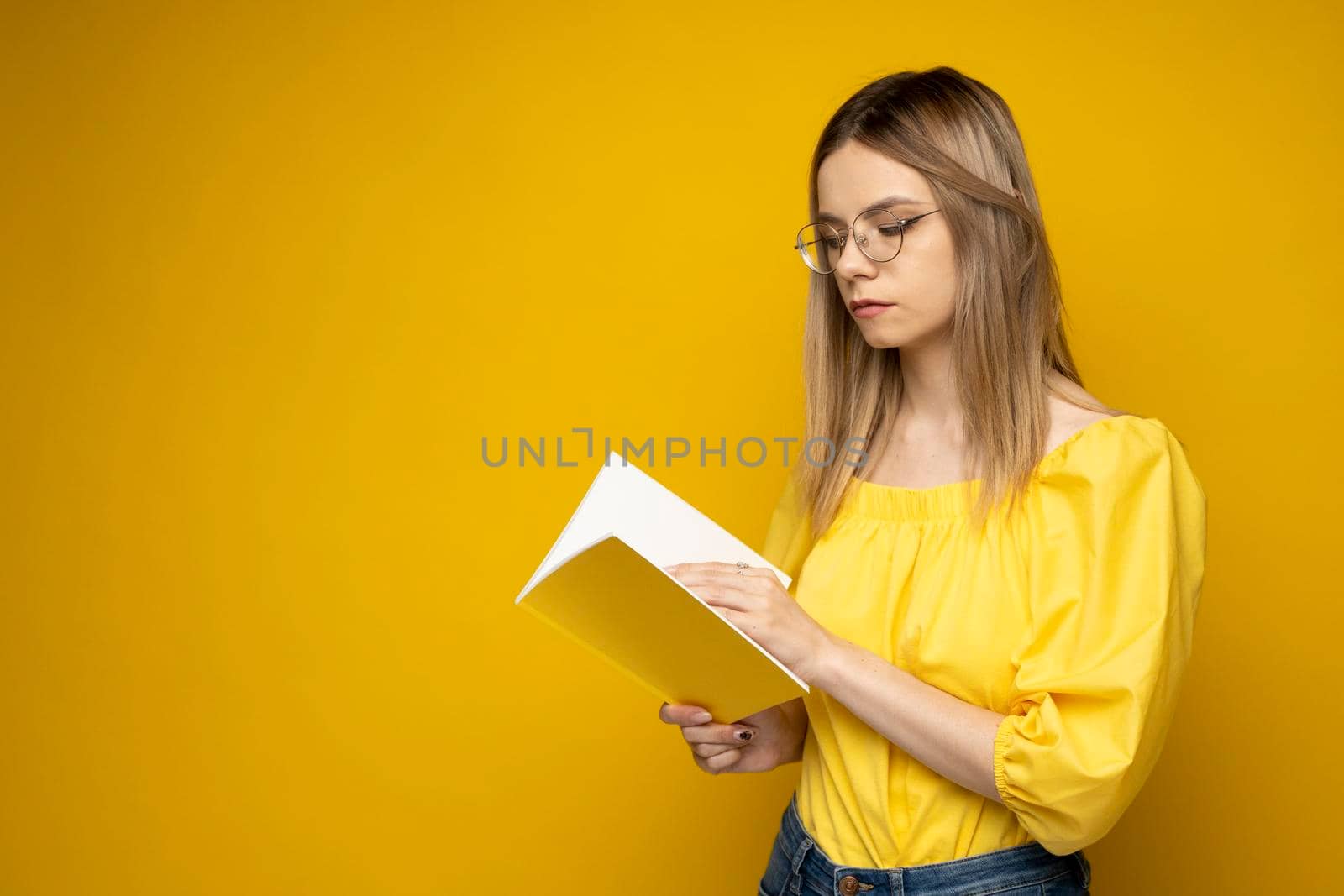 Beautiful smart young girl holding and reading book isolated on the yellow background. Portrait of attractive woman in a yellow blouse and wearing glasses reading book. Education, studying, knowledge. by vovsht