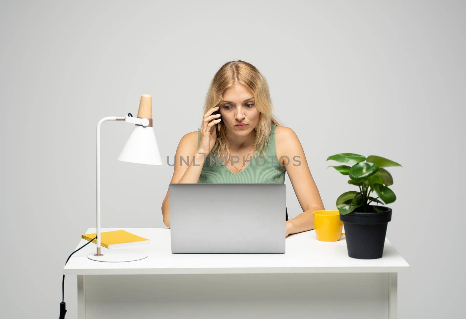 Young attractive business woman with a blond hair sitting at the table and working on a laptop and talking with a client on the phone. Young cheerful student girl talking with a smartphone. Studying. by vovsht