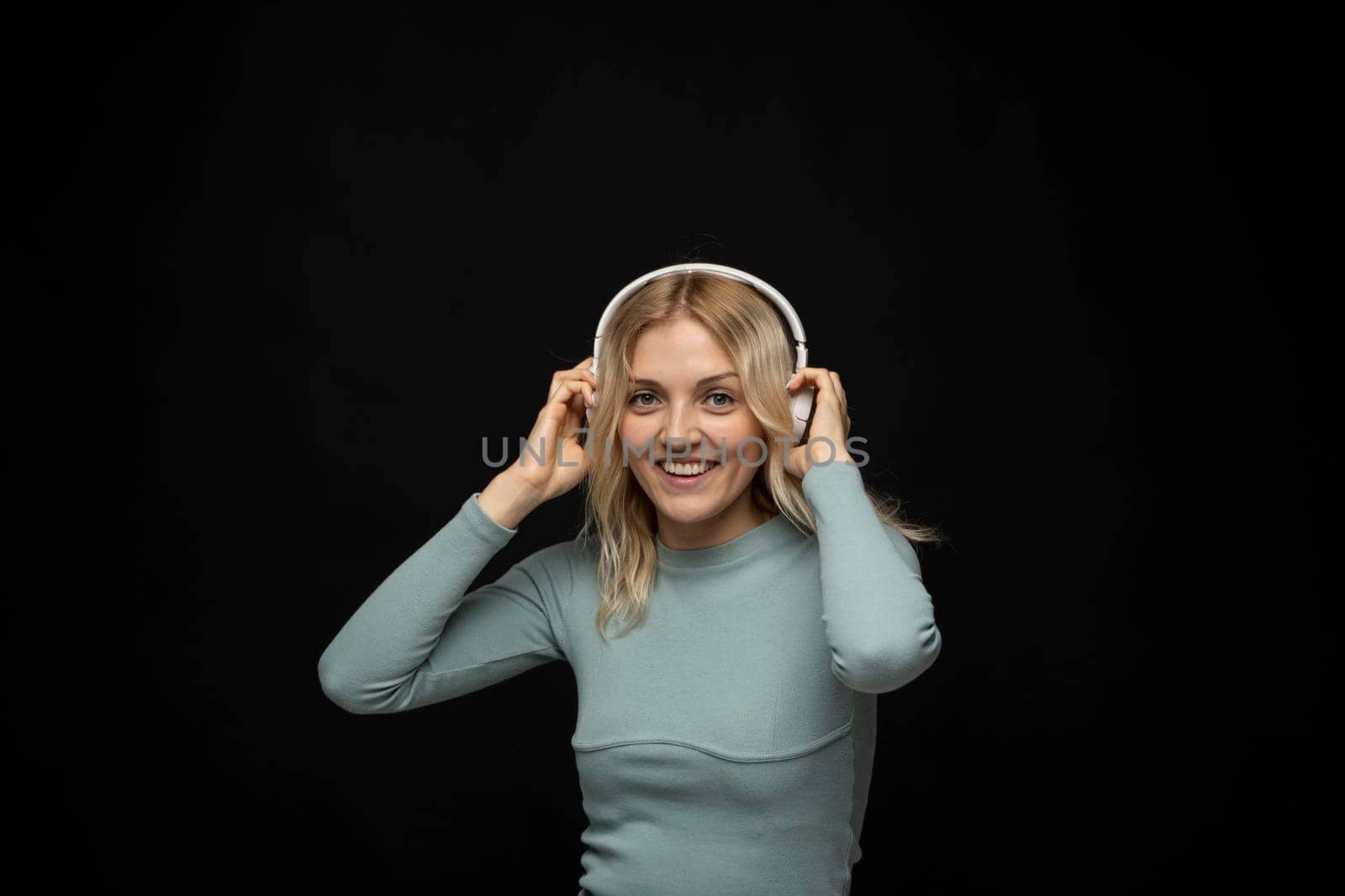 Beautiful attractive young blond woman wearing blue t-shirt and glasses in white headphones listening music and smiling on black background in studio. Relaxing and enjoying. Lifestyle