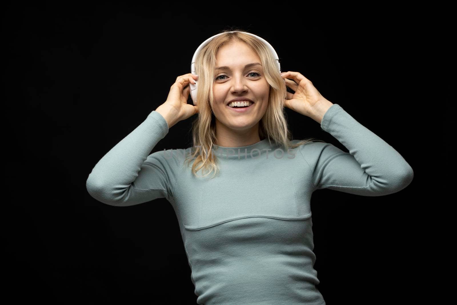 Beautiful attractive young blond woman wearing blue t-shirt and glasses in white headphones listening music and smiling on black background in studio. Relaxing and enjoying. Lifestyle