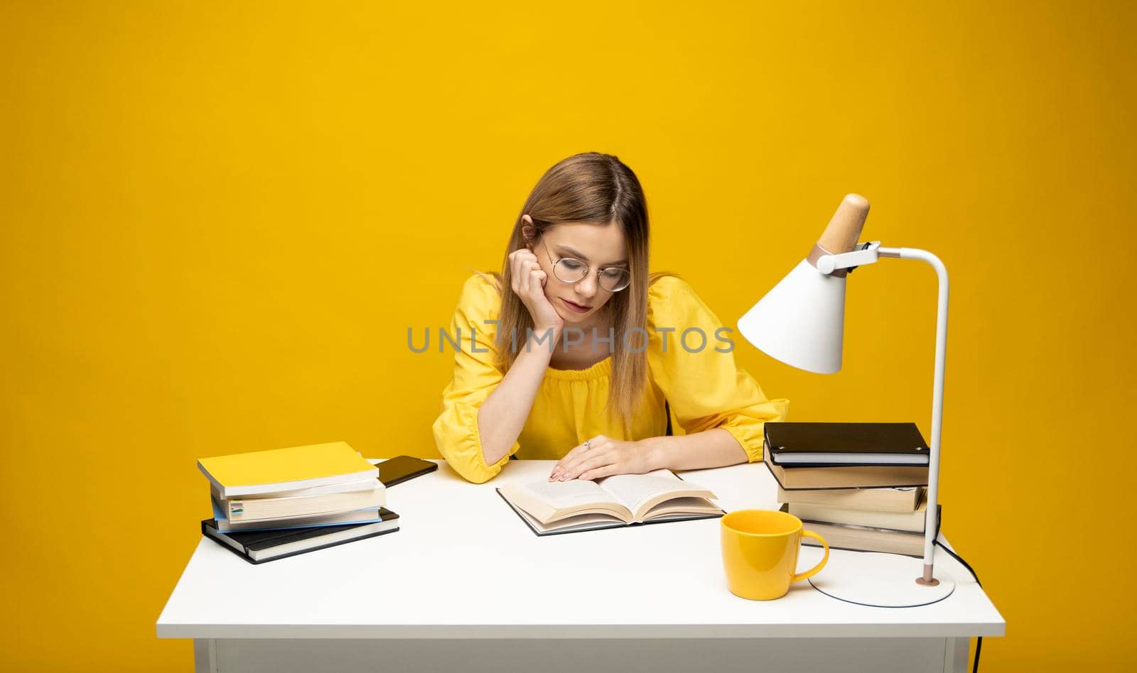 Tired young student woman in yellow casual clothes sitting at the table reading the book in library of university or college. Sitting and reading on yellow background. Studying