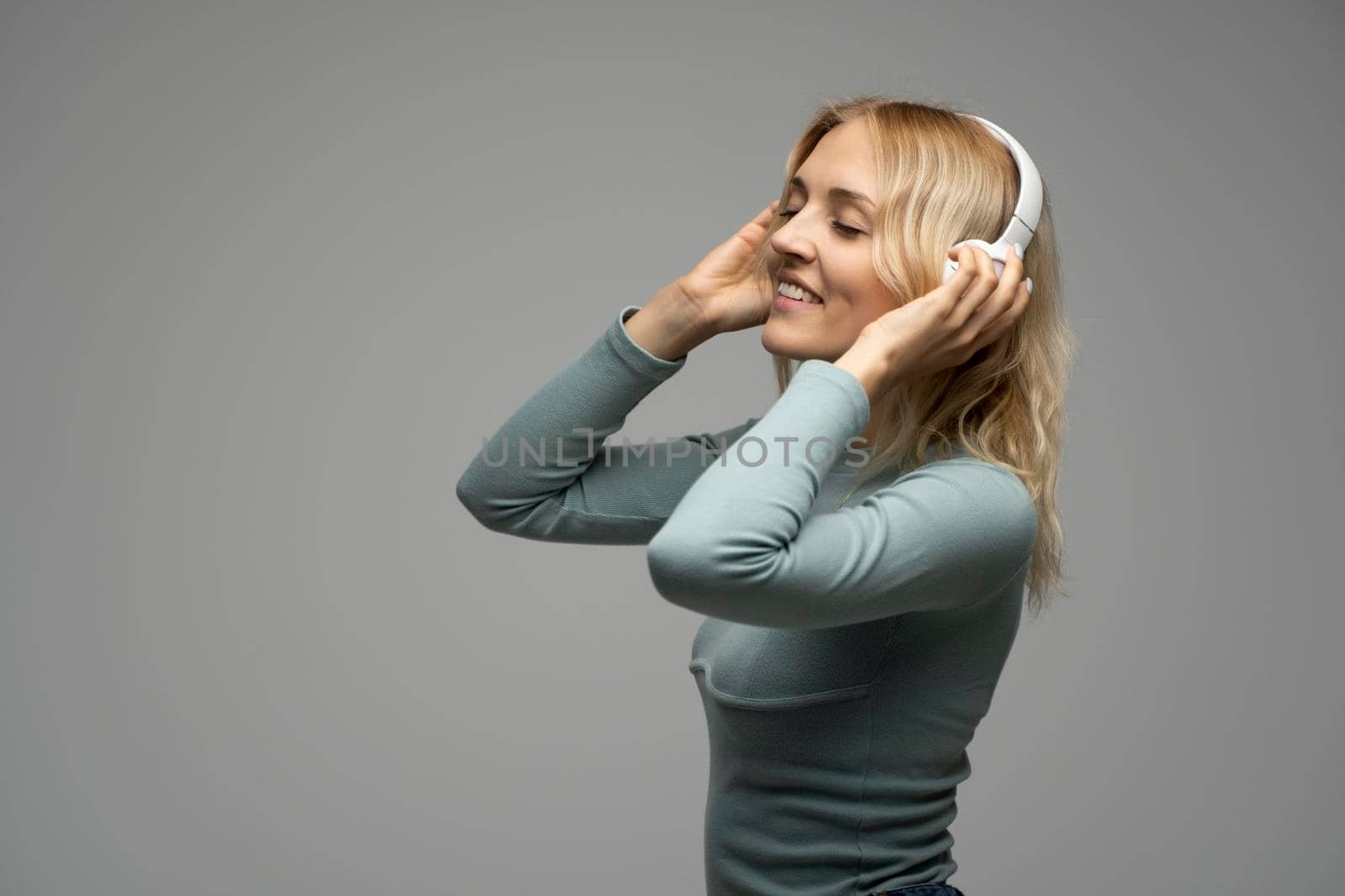 Beautiful attractive young blond woman wearing blue t-shirt and glasses in white headphones listening music and smiling on grey background in studio. Relaxing and enjoying. Lifestyle