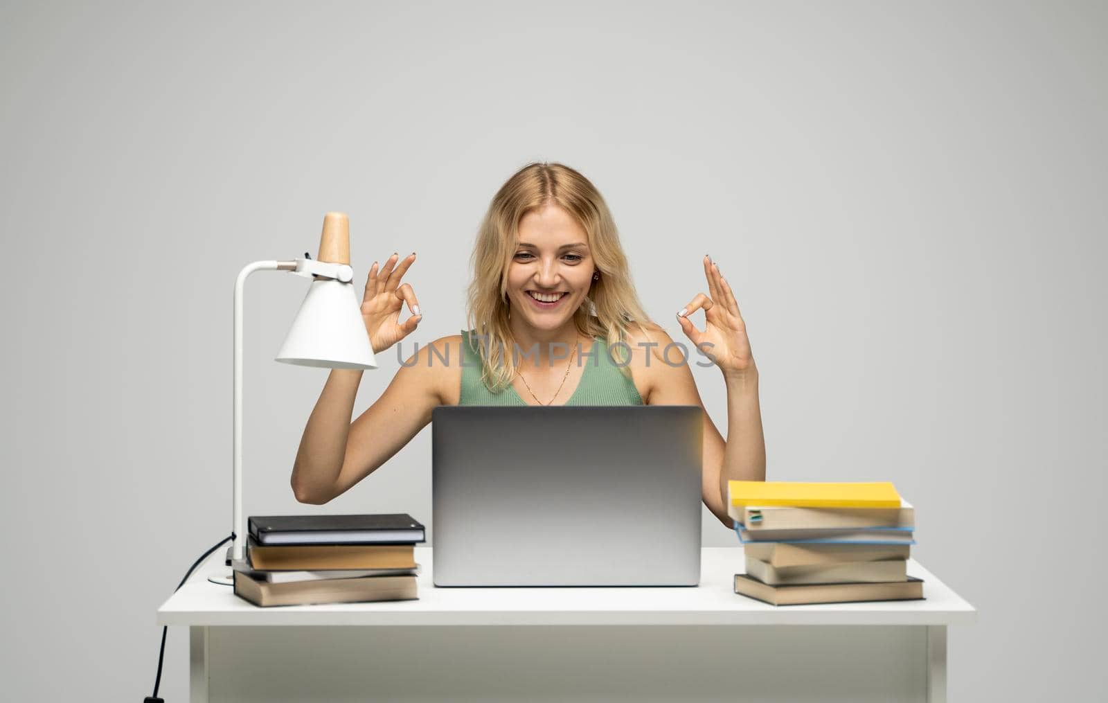 Student girl showing ok gesture with both hands, happy with completed work, smiling and looking at the camera while working on laptop computer sitting in front of big piles of books. by vovsht