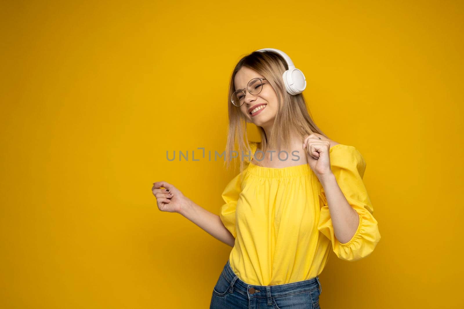 Blonde woman in a glasses and yellow shirt listening music with a headphones. Teenager girl dancing against yellow background. by vovsht