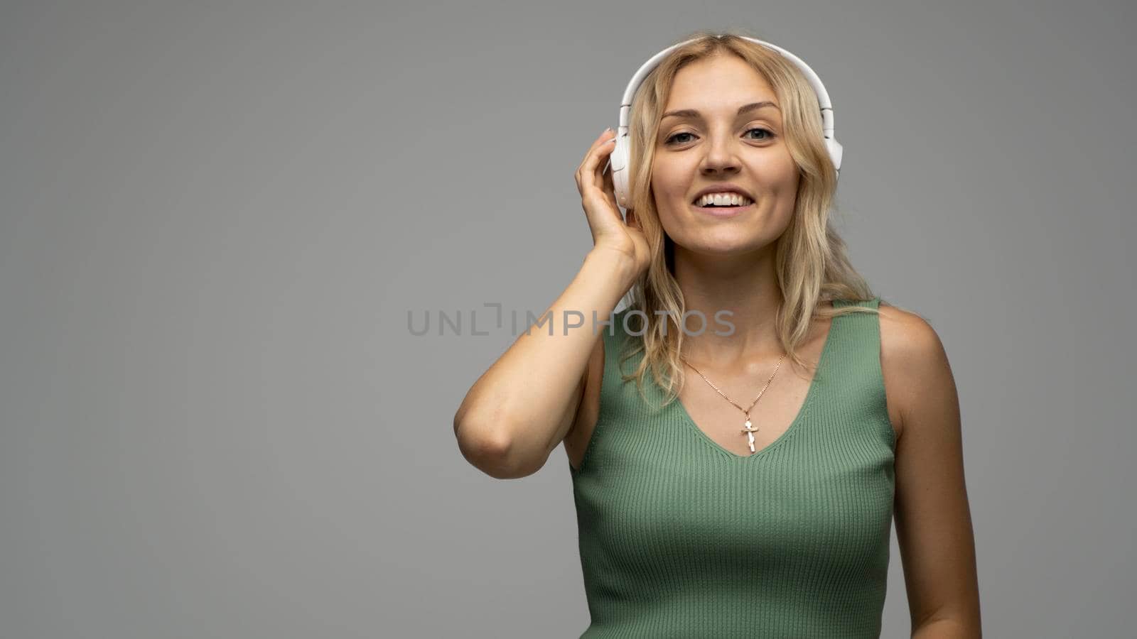 Beautiful attractive young blond woman wearing green t-shirt and glasses in white headphones listening music and smiling on grey background in studio. Relaxing and enjoying. Lifestyle. by vovsht