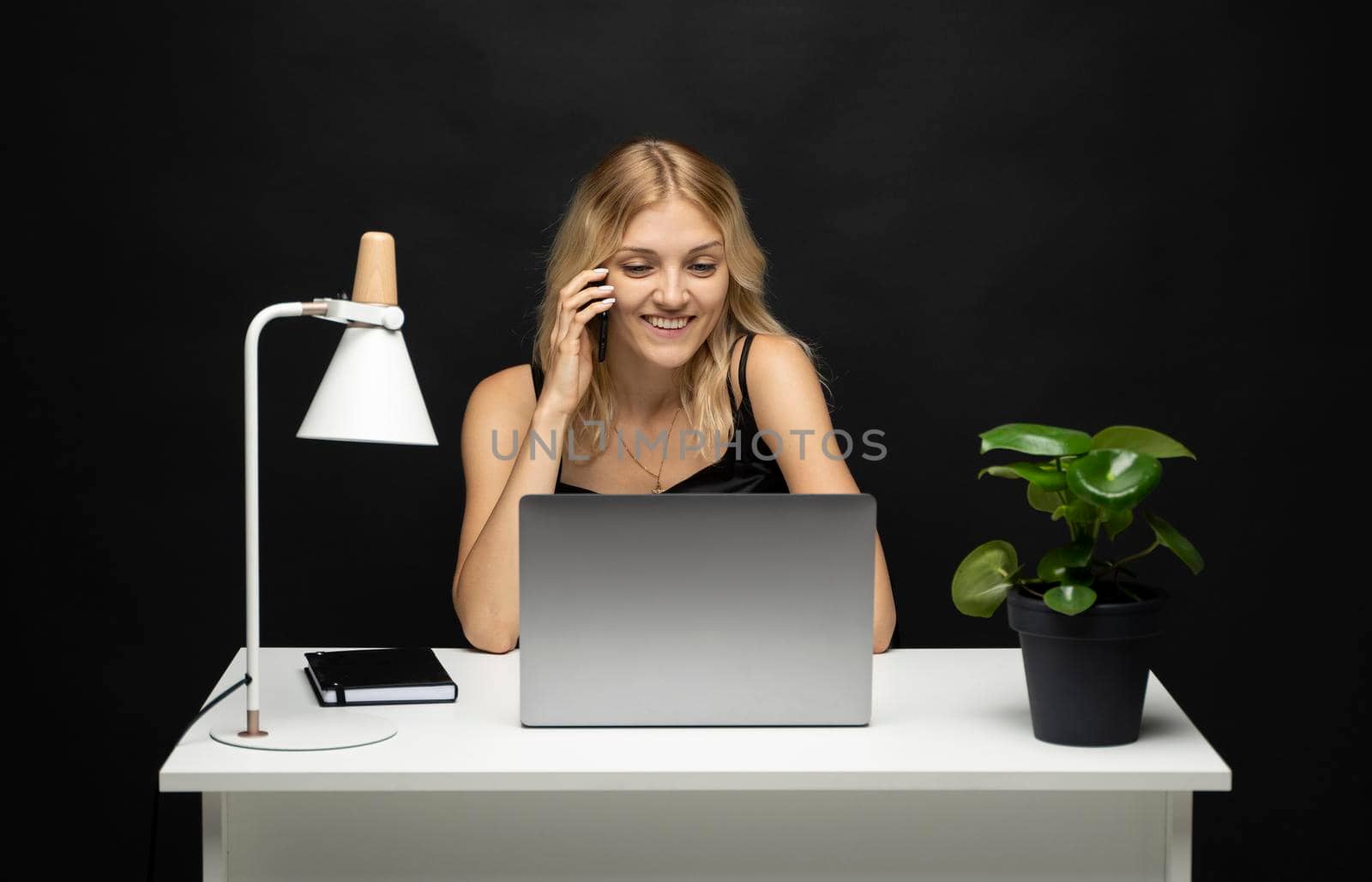 Young attractive business woman with a blond hair sitting at the table and working on a laptop and talking with a client on the phone. Young cheerful student girl talking with a smartphone. Studying. by vovsht