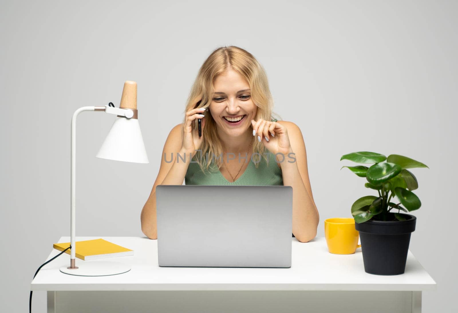 Young attractive business woman with a blond hair sitting at the table and working on a laptop and talking with a client on the phone. Young cheerful student girl talking with a smartphone. Studying. by vovsht