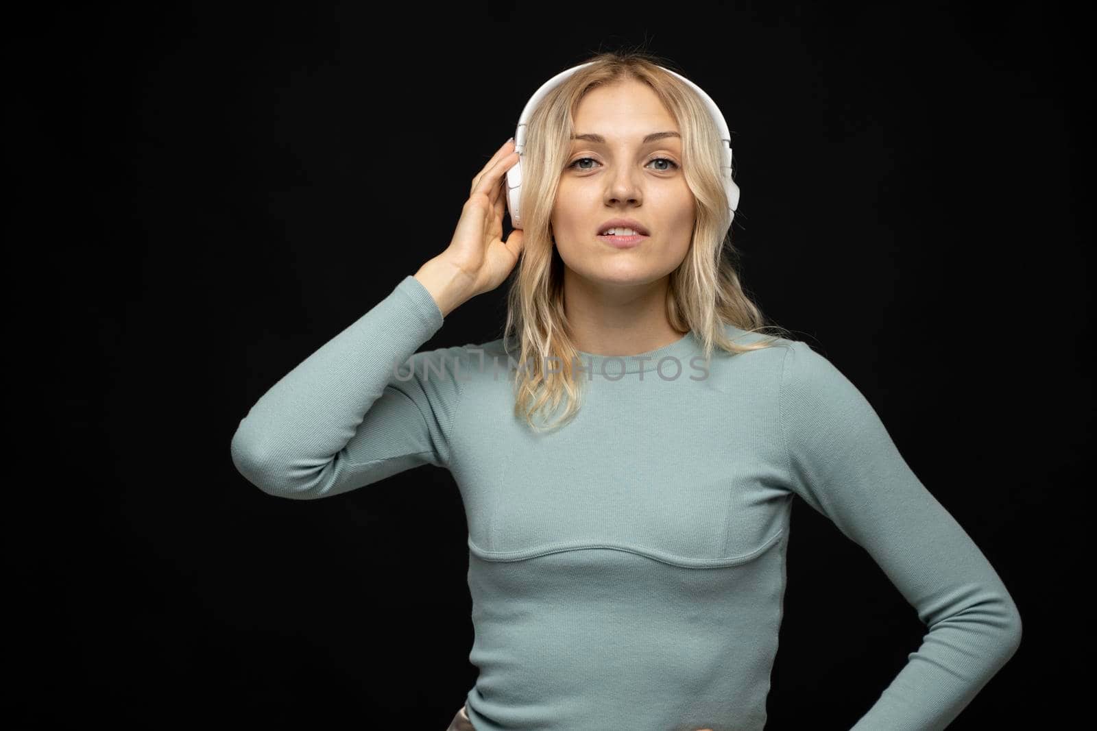 Beautiful attractive young blond woman wearing blue t-shirt and glasses in white headphones listening music and smiling on black background in studio. Relaxing and enjoying. Lifestyle. by vovsht