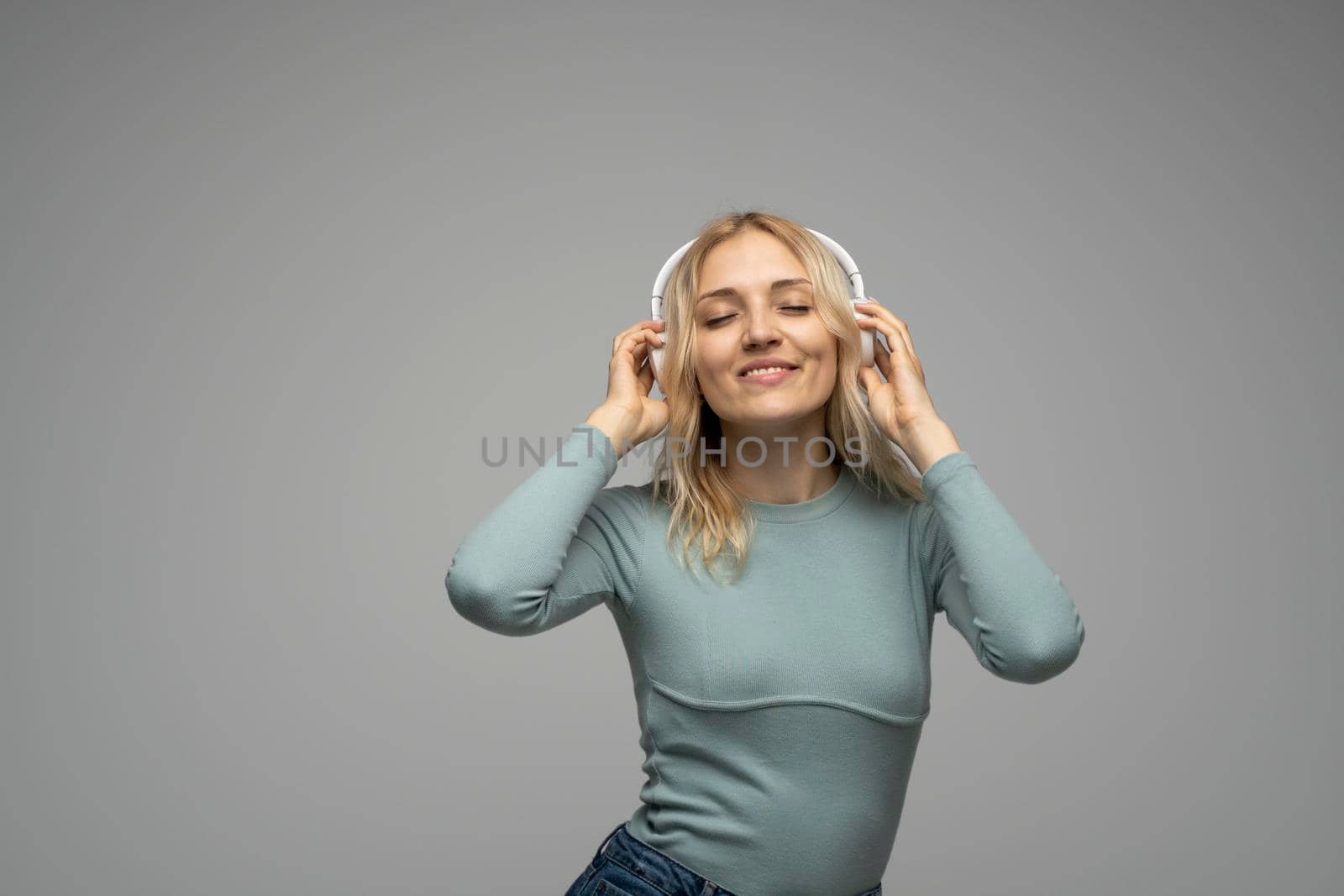 Beautiful attractive young blond woman wearing blue t-shirt and glasses in white headphones listening music and smiling on grey background in studio. Relaxing and enjoying. Lifestyle