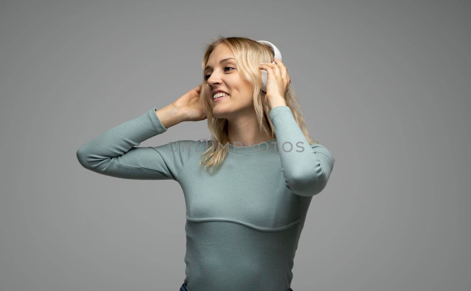 Beautiful attractive young blond woman wearing blue t-shirt and glasses in white headphones listening music and smiling on grey background in studio. Relaxing and enjoying. Lifestyle. by vovsht