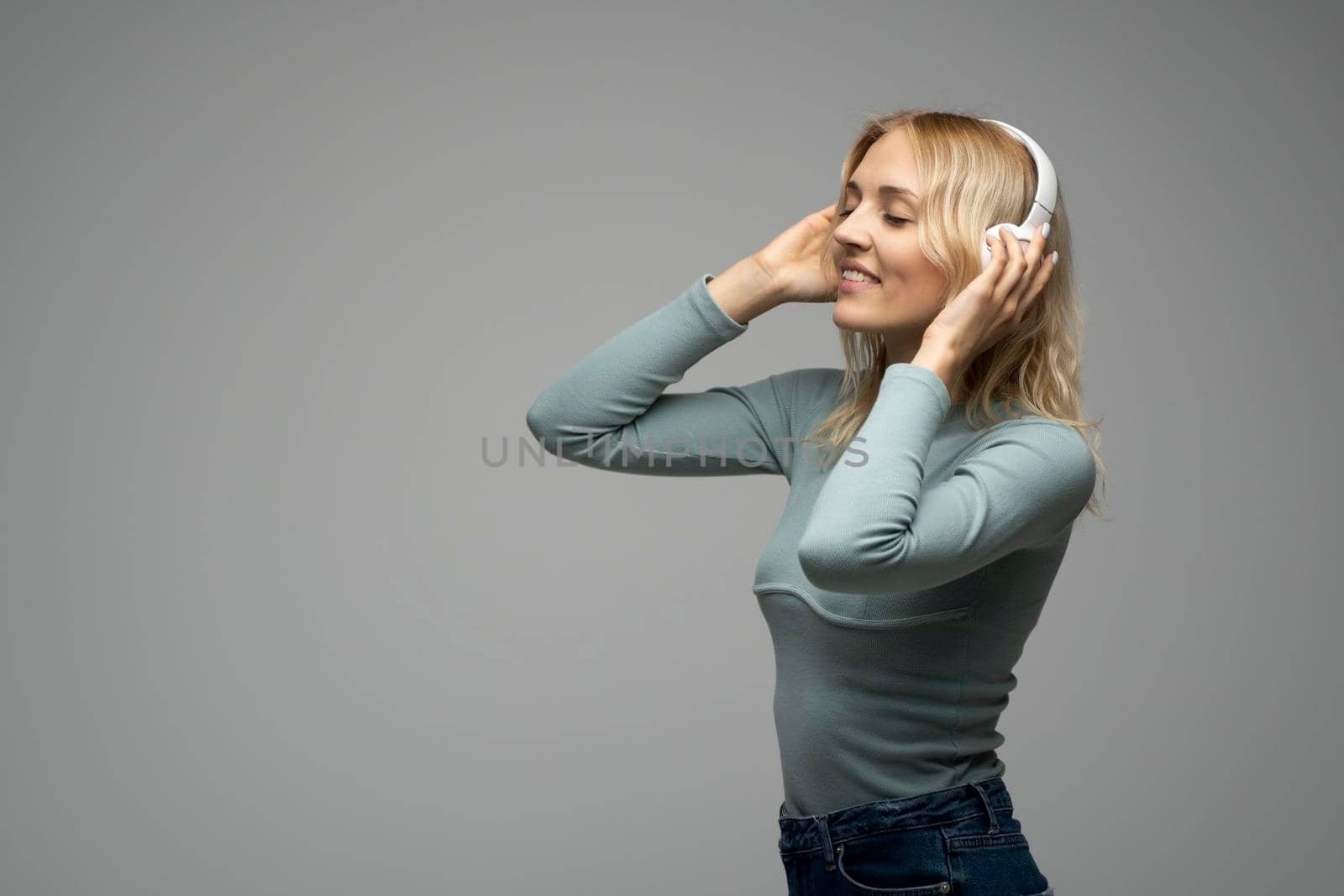 Beautiful attractive young blond woman wearing blue t-shirt and glasses in white headphones listening music and smiling on grey background in studio. Relaxing and enjoying. Lifestyle