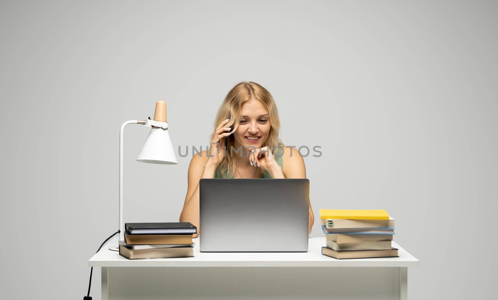 Young attractive business woman with a blond hair sitting at the table and working on a laptop and talking with a client on the phone. Young cheerful student girl talking with a smartphone. Studying. by vovsht