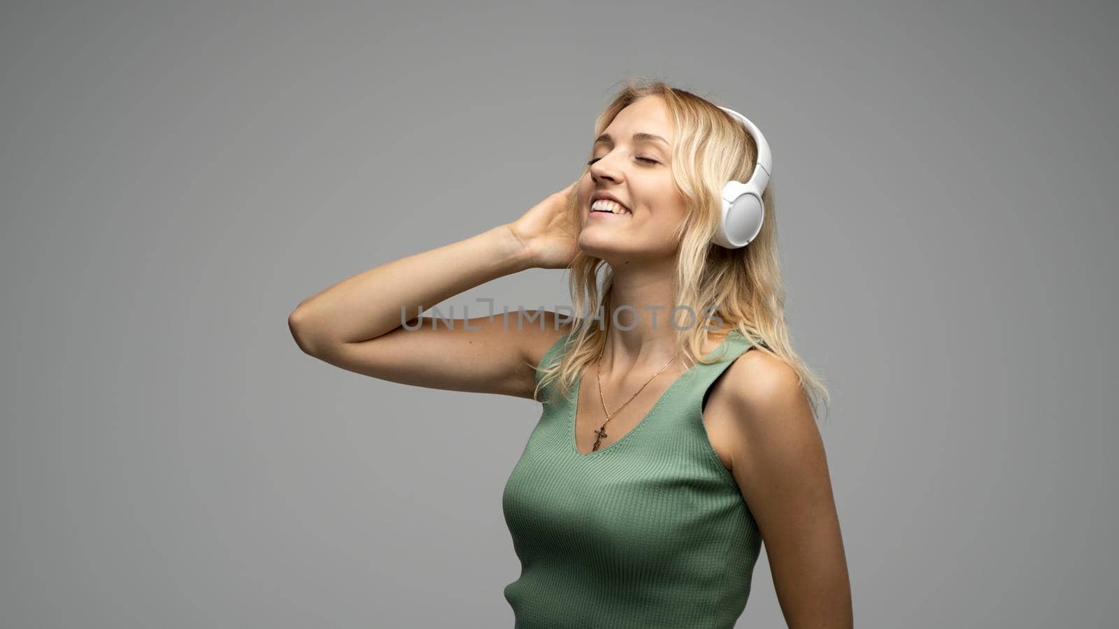 Blonde girl with white headphones listening to music with closed eyes on grey background in studio. She wears grey T-shirt