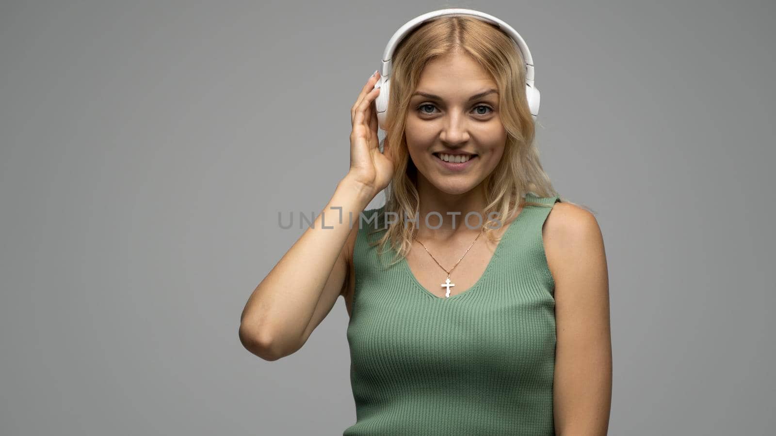 Beautiful attractive young blond woman wearing green t-shirt and glasses in white headphones listening music and smiling on grey background in studio. Relaxing and enjoying. Lifestyle. by vovsht