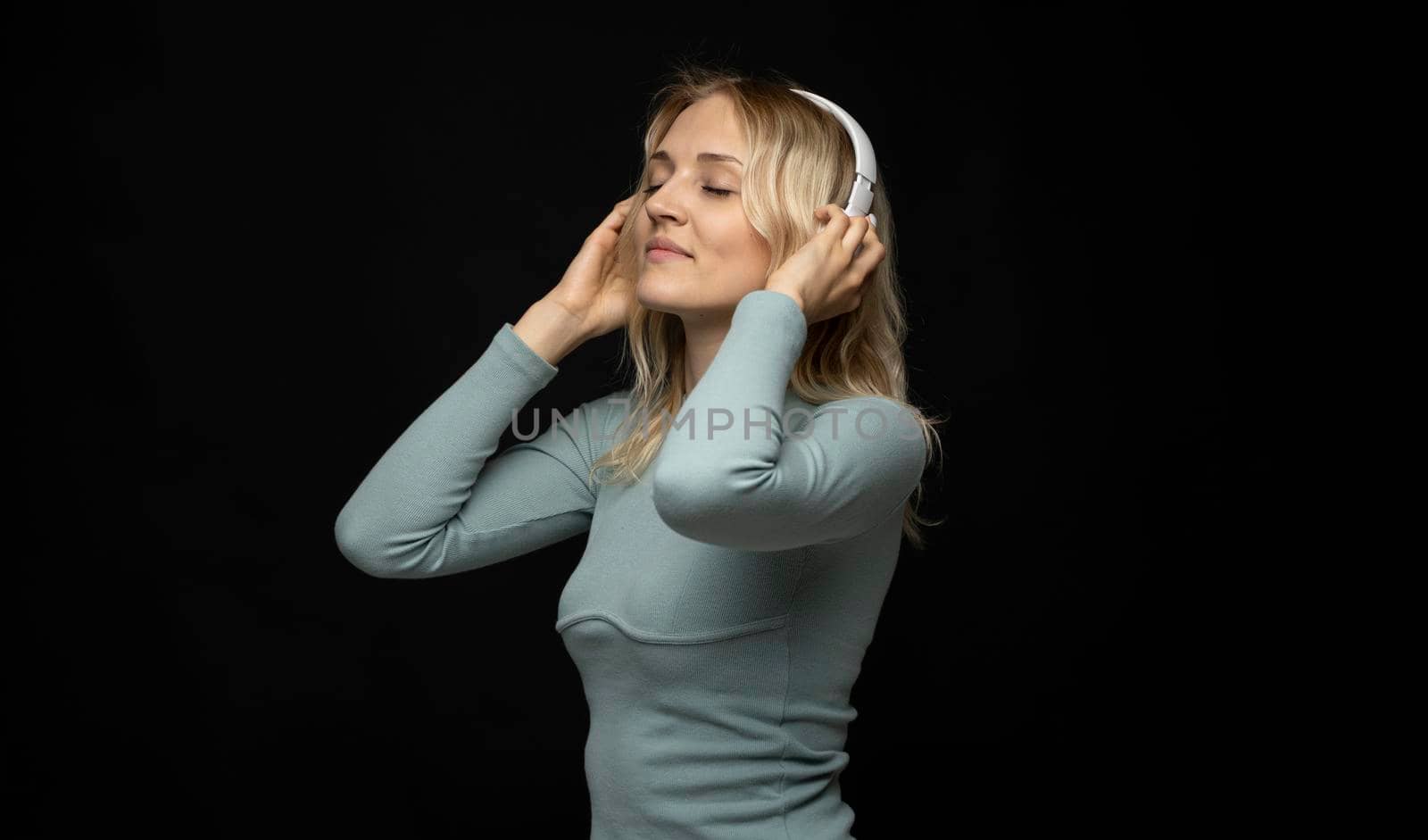 Beautiful attractive young blond woman wearing blue t-shirt and glasses in white headphones listening music and smiling on black background in studio. Relaxing and enjoying. Lifestyle. by vovsht