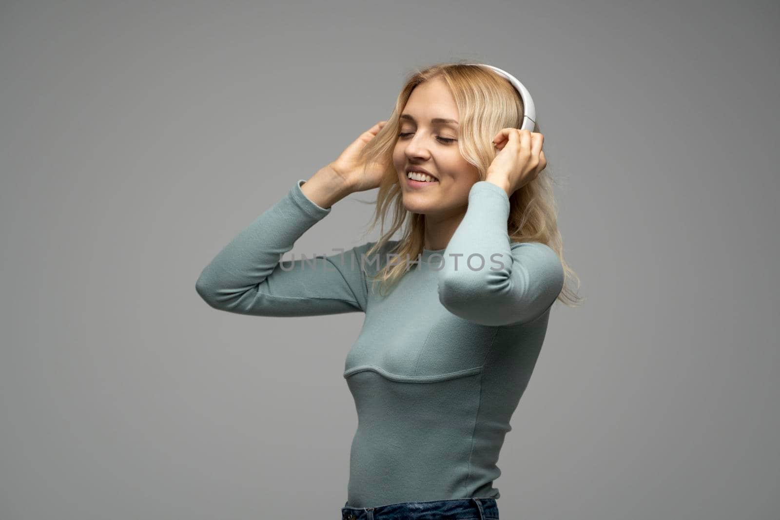 Beautiful attractive young blond woman wearing blue t-shirt and glasses in white headphones listening music and smiling on grey background in studio. Relaxing and enjoying. Lifestyle. by vovsht