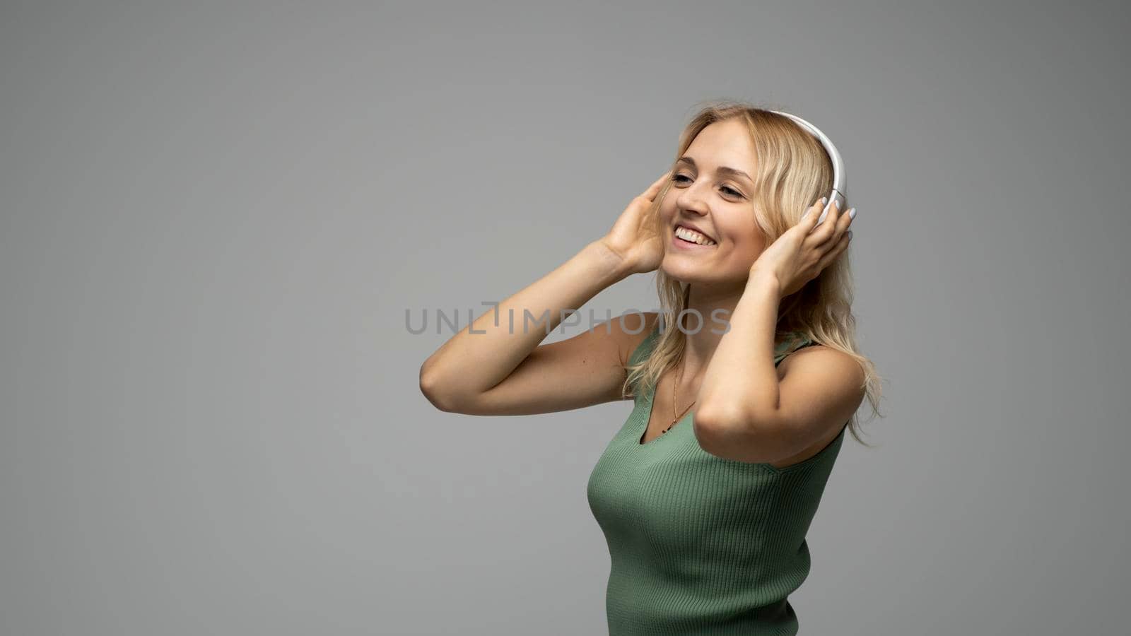 Beautiful attractive young blond woman wearing green t-shirt and glasses in white headphones listening music and smiling on grey background in studio. Relaxing and enjoying. Lifestyle. by vovsht