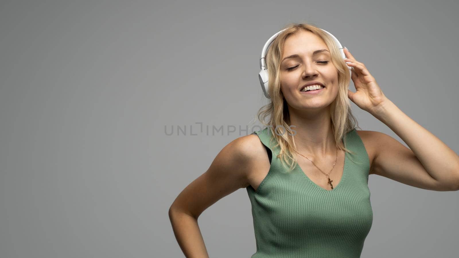 Beautiful attractive young blond woman wearing green t-shirt and glasses in white headphones listening music and smiling on grey background in studio. Relaxing and enjoying. Lifestyle. by vovsht