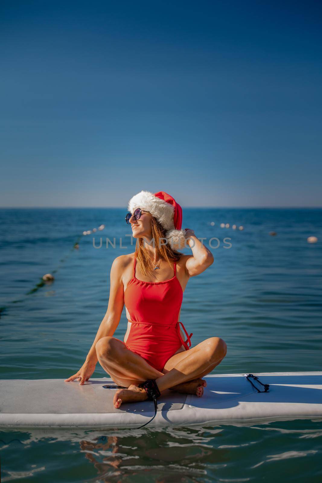 A woman in a red swimsuit and a Santa hat with glasses sits on a SUP board at the sea. He looks away and smiles by Matiunina