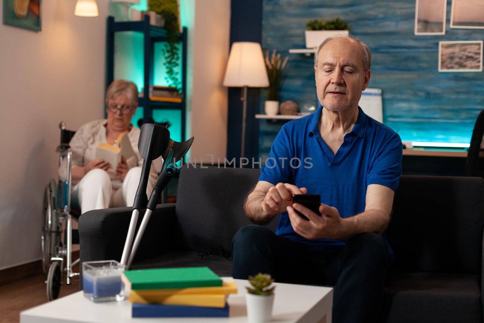Aged caucasian man using smartphone on living room sofa. Senior adult holding modern device with technology looking at screen while old woman in wheelchair sitting in background