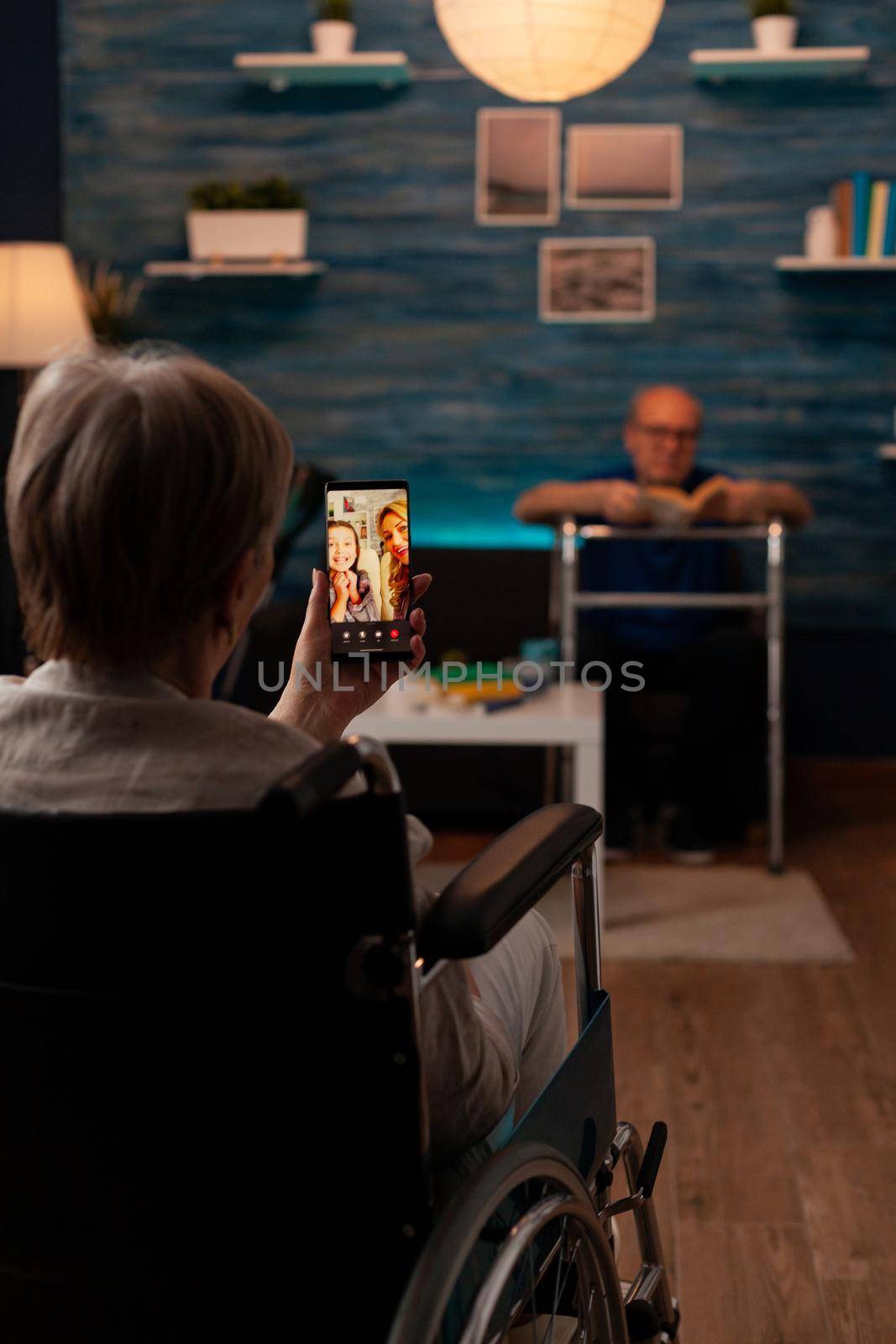 Aged disabled woman holding smartphone for video call conference with relatives while sitting in living room. Senior pensioner talking to daughter and niece with online internet in wheelchair