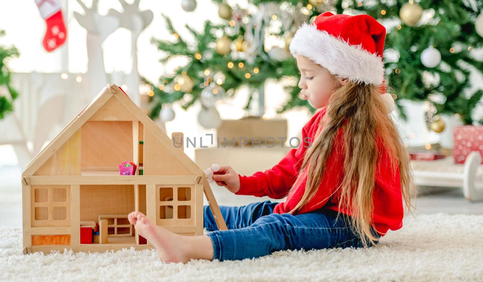 Little girl child wearing Santa hat in Christmas time sitting on floor and playing with wooden toy house at decorated New Year home with festive tree. Cute kid in Xmas time