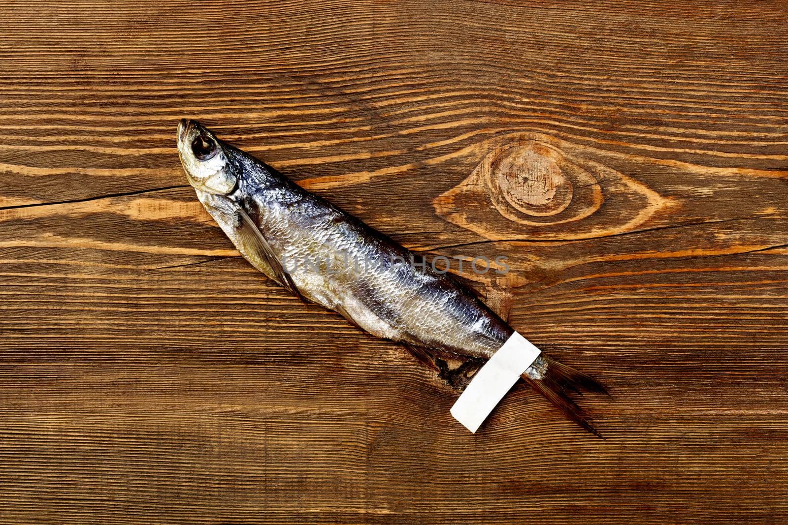 One jerked or dried salted Baltic herring with paper label on tail lying on rustic wooden table. Traditional way of preserving fish