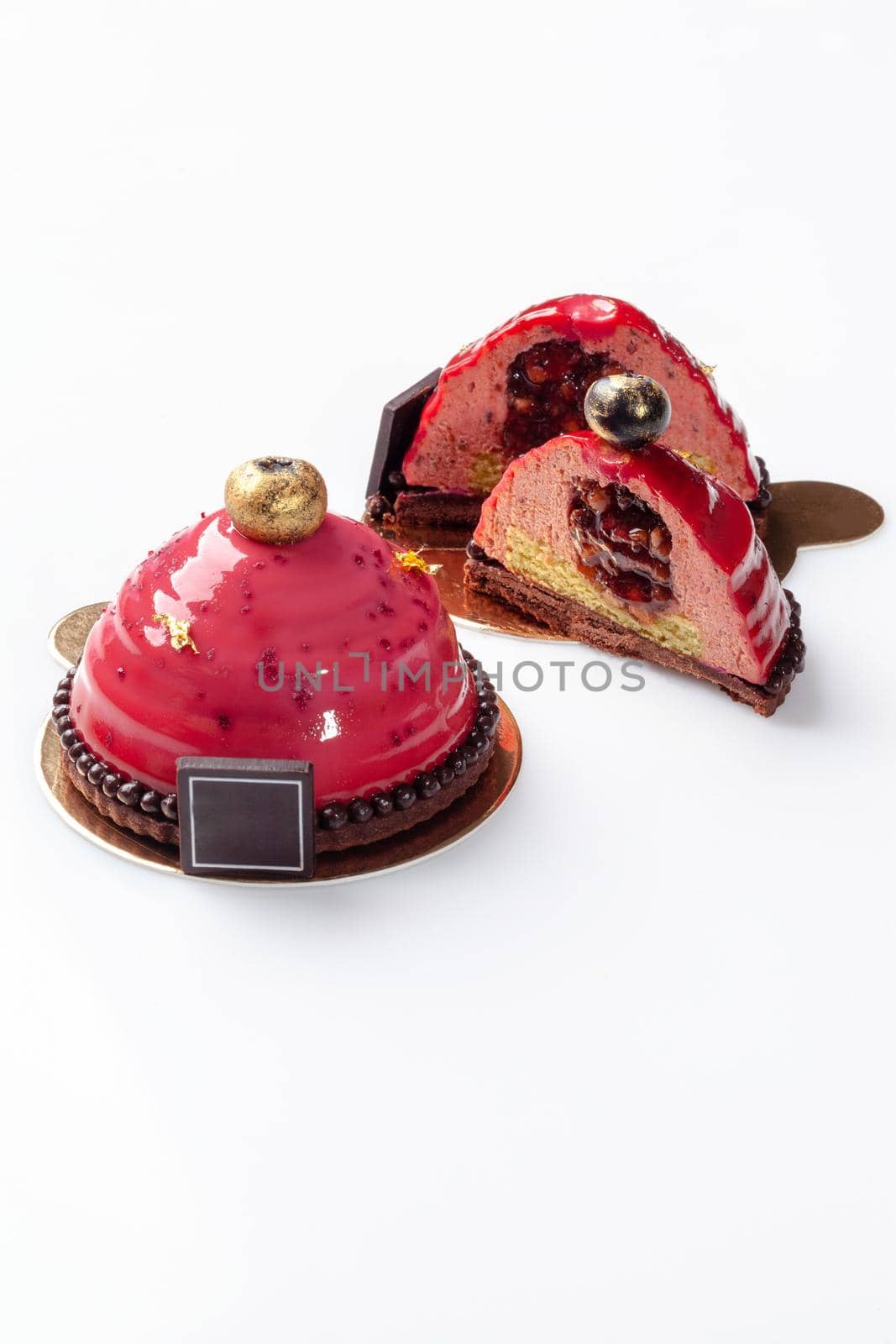 Tempting dessert, whole and cut in half, of airy plum mousse on chocolate and nut shortcrust base with fruit filling topped with pink glaze and decorated with edible gold on white background