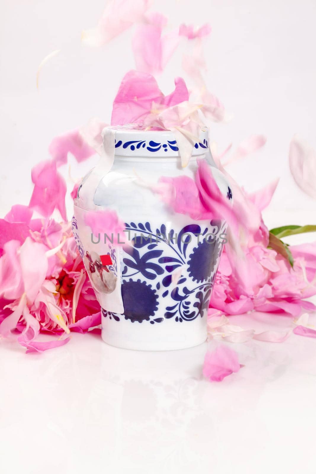 Decorative jar with blue floral patterns sprinkled with pink peony petals on white background. Packaging for storing food or cosmetics