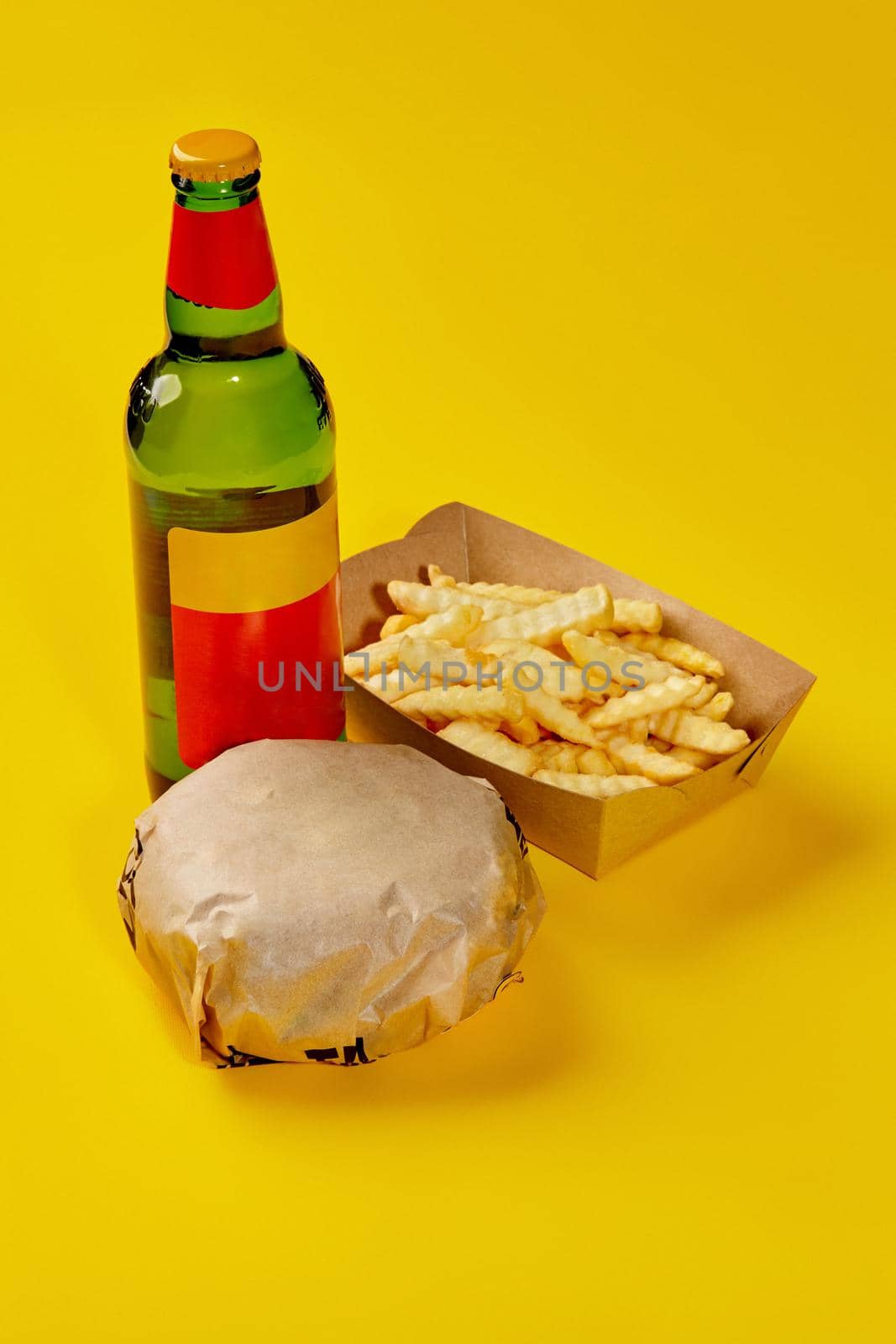 Fresh hamburger in paper wrapping, crispy French fries in carton box and glass bottle of drink isolated on yellow background. Set from fast food restaurant menu. Popular snacks concept