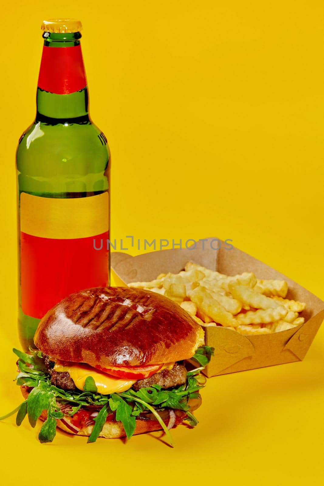 Delicious burger in white wheat glossy bun with veal patty, cheese, tomatoes, onion and fresh arugula leaves served with crispy fries in paper box and drink in bottle isolated on yellow background