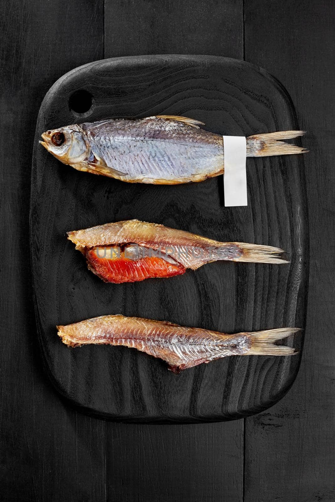 Whole salted air-dried roach fish with label on tail and two peeled fish with caviar served on black wooden board. Popular savory snack