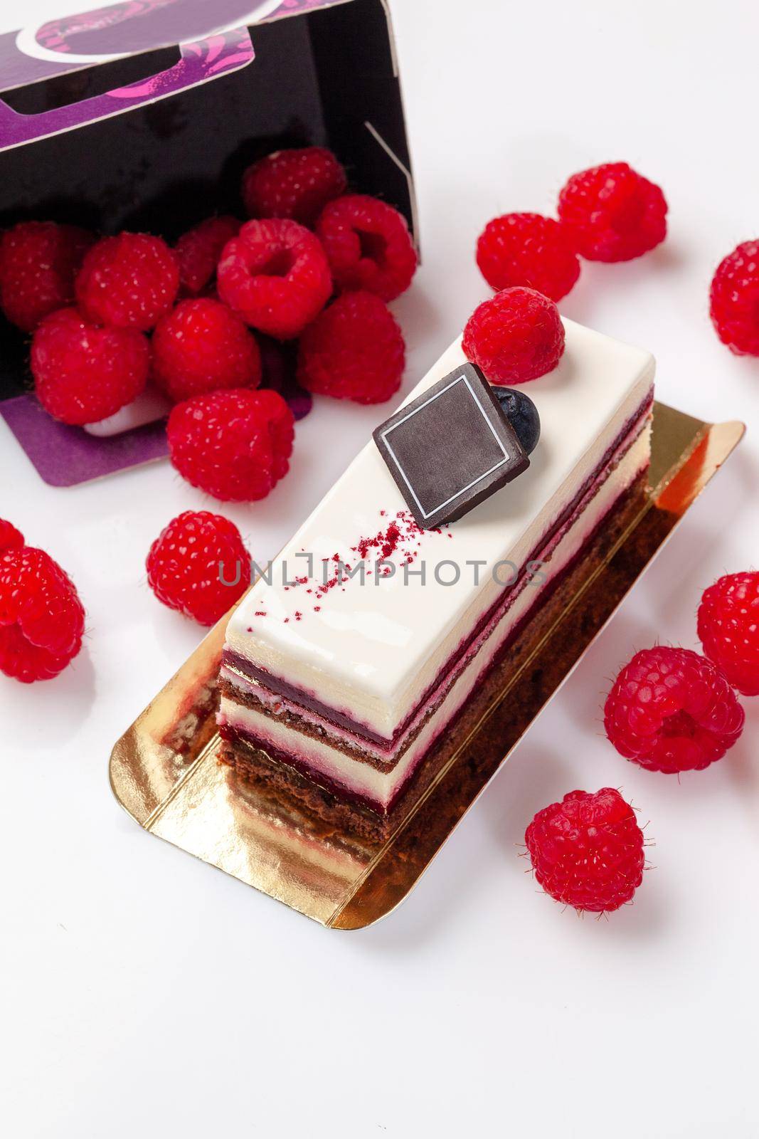 Chocolate sponge cake slice with layers of whipped cheese cream and aromatic raspberry confit garnished with fresh ripe berries on white surface. Delicious sweet pastry
