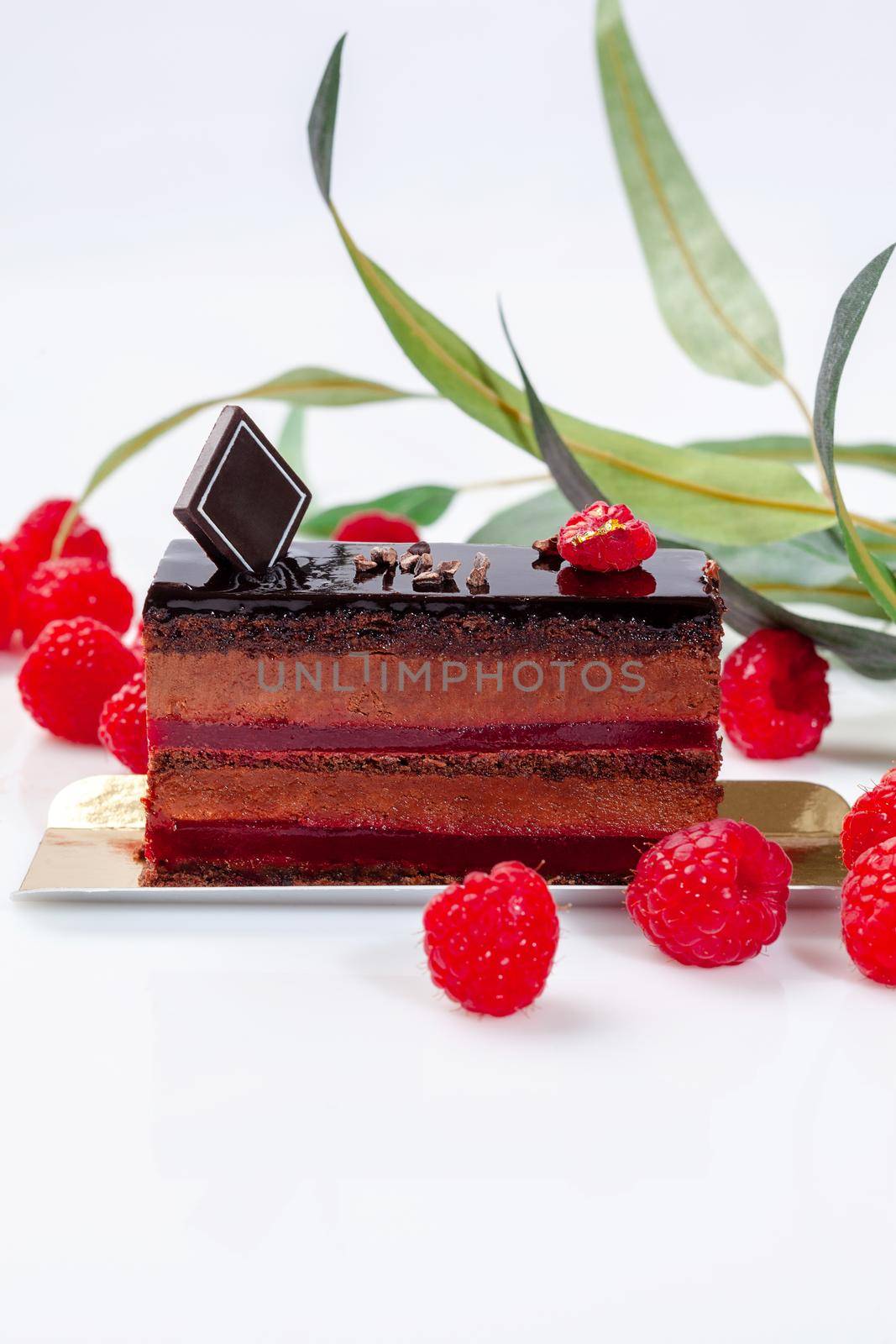 Delicious moist chocolate sponge cake with airy chocolate mousse and raspberry confit interlayer topped with glaze and garnished with fresh berries on white background