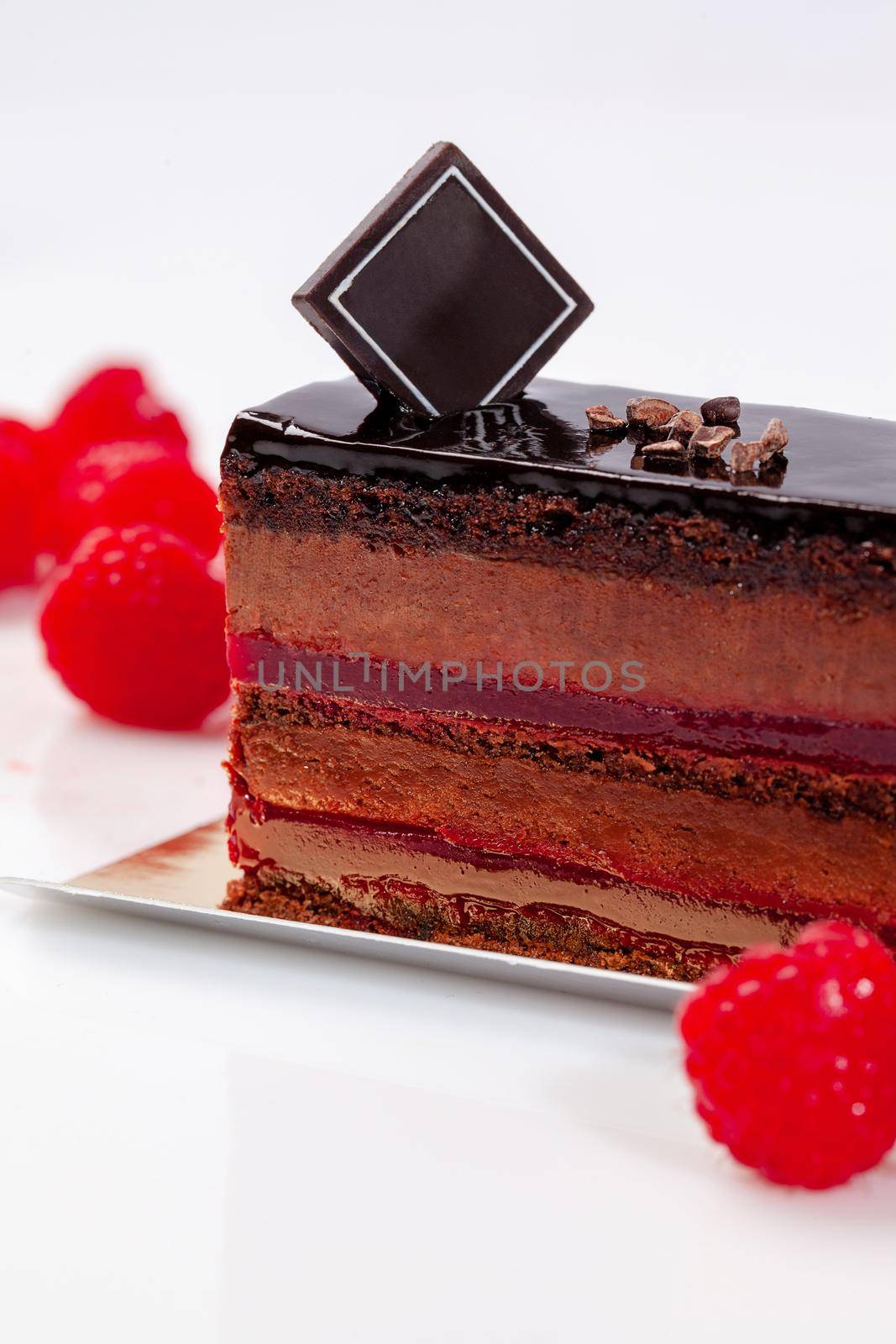 Slice of tempting chocolate cake with delicate airy mousse and raspberry jelly decorated with cocoa nibs and fresh berries on white background