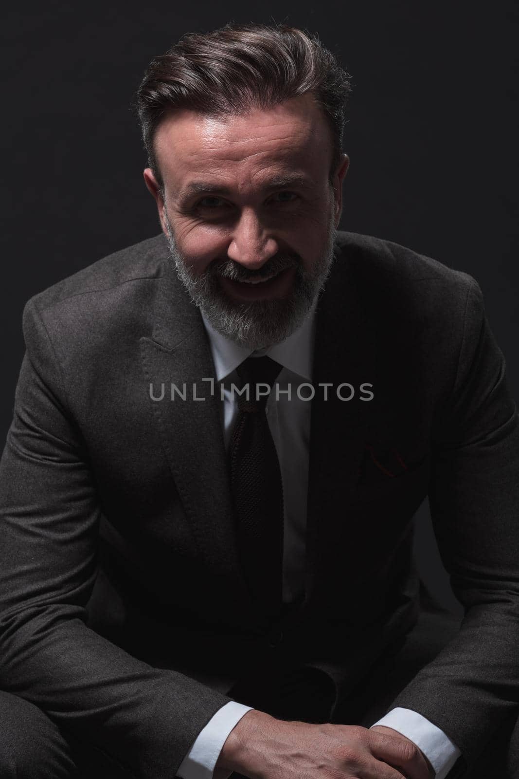 Portrait of adult businessman wearing trendy suit and sitting in modern studio on stylish chair against the black background by dotshock