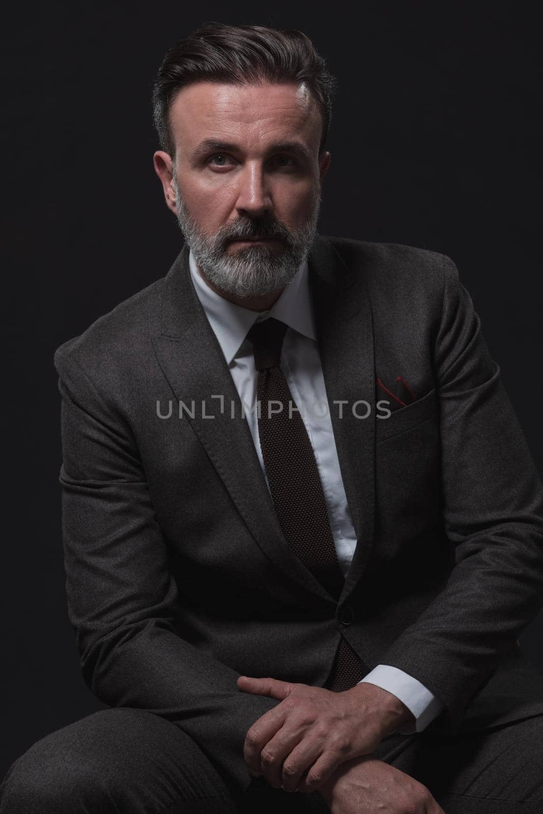 Portrait of adult businessman wearing trendy suit and sitting in modern studio on stylish chair against the black background by dotshock