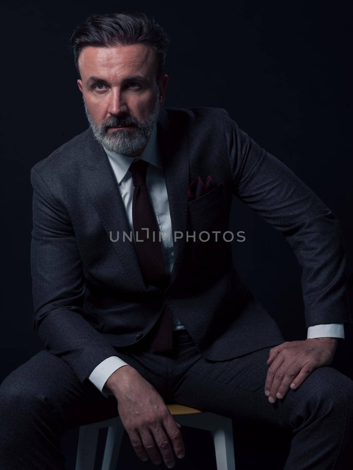 Portrait of adult businessman wearing trendy suit and sitting in modern studio on stylish chair against the black background. Horizontal mockup. High quality photo