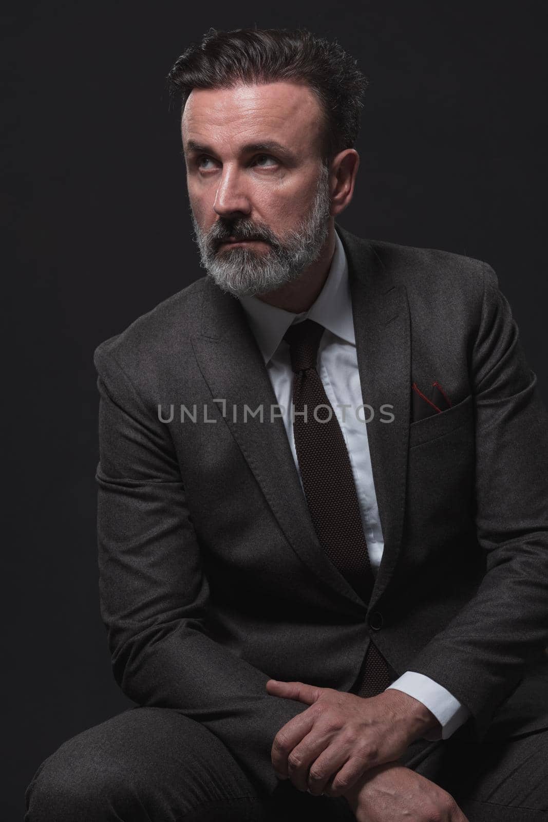 Portrait of adult businessman wearing trendy suit and sitting in modern studio on stylish chair against the black background. Horizontal mockup. High quality photo