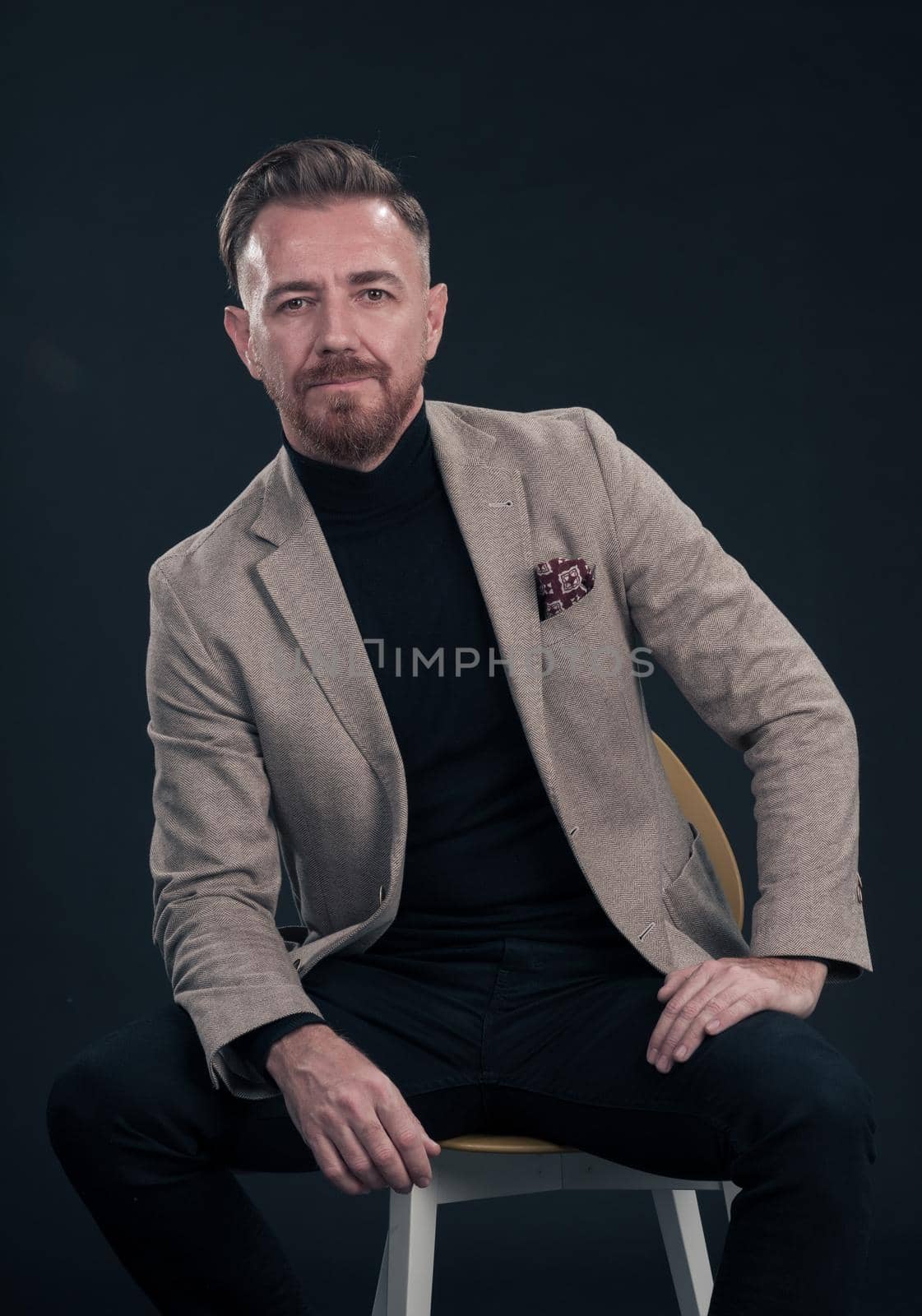 Portrait of adult businessman wearing trendy suit and sitting in modern studio on stylish chair against the black background. Horizontal mockup. High quality photo