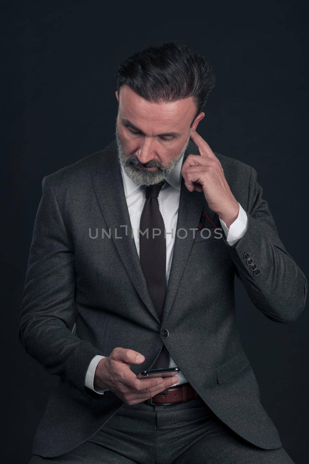 Chat with the client. Good looking young man in full suit using his smart phone and smiling while standing against dark grey background. High quality photo