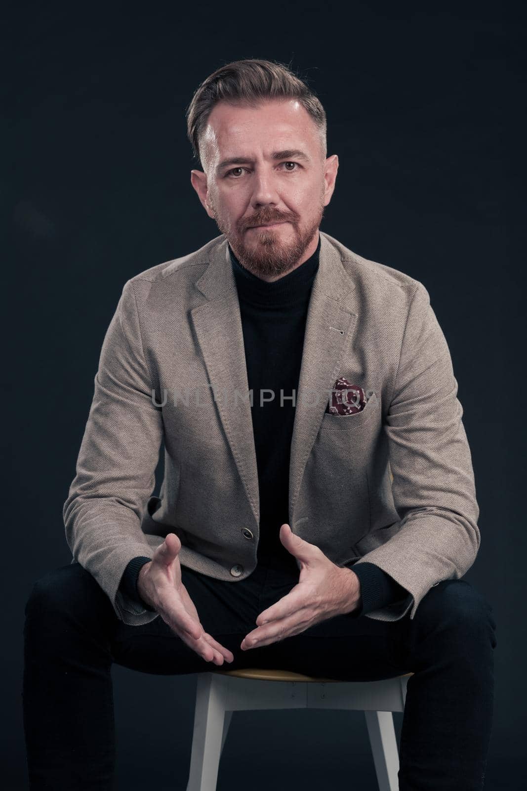 Portrait of adult businessman wearing trendy suit and sitting in modern studio on stylish chair against the black background. Horizontal mockup. High quality photo