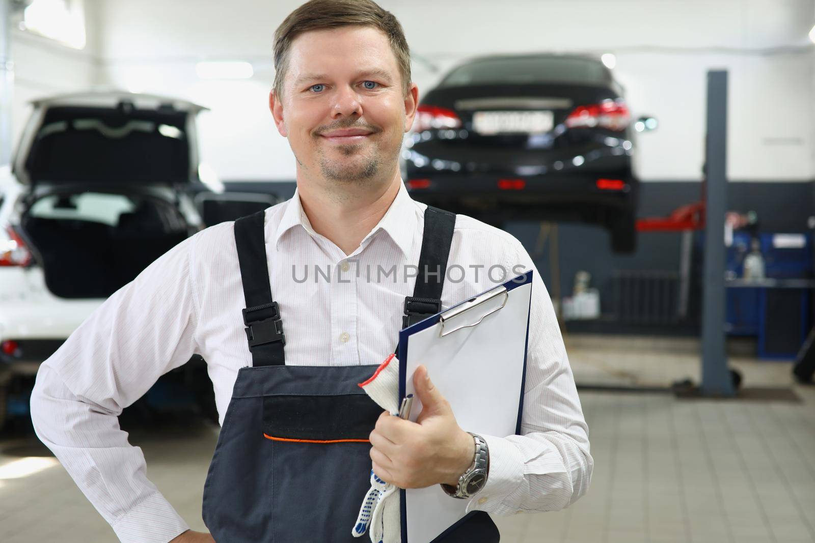 Portrait of qualified maintenance centre worker in special uniform posing on cars background. Mechanic male propose high service. Pit stop, garage concept