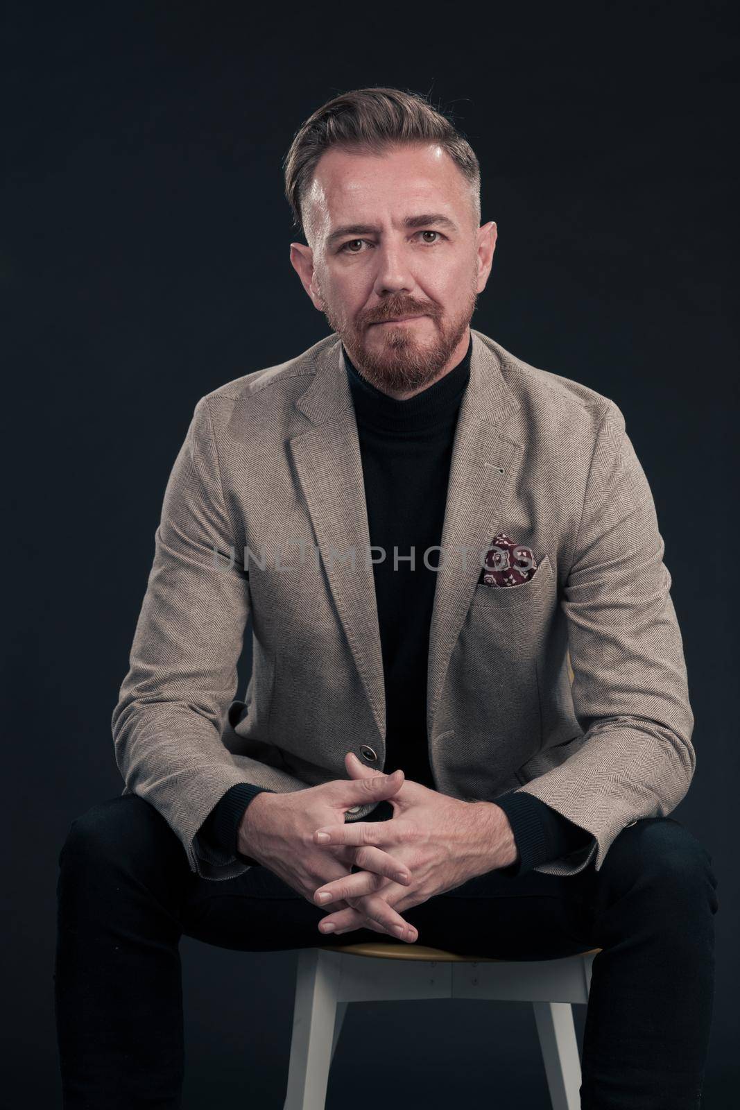 Portrait of adult businessman wearing trendy suit and sitting in modern studio on stylish chair against the black background. Horizontal mockup. High quality photo