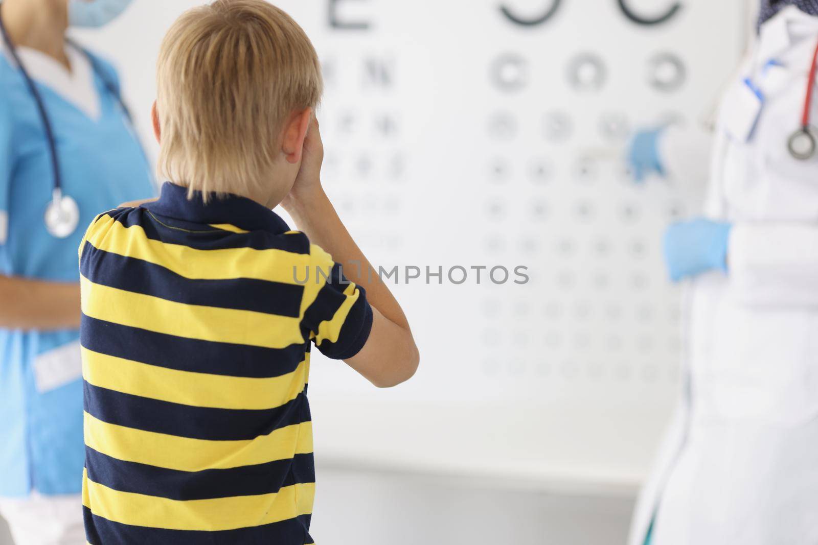 Portrait of little boy on appointment at oculist office, check sight closing eye and say out loud letter on board. Childhood, ophthalmology, health concept