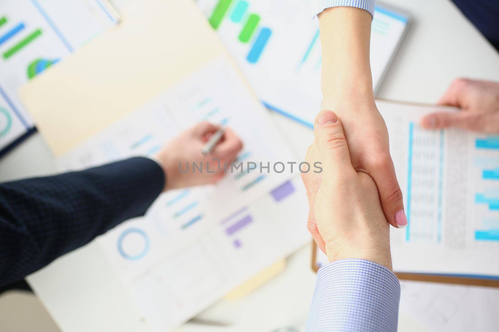 Top view of partners shake hands over business papers on desk. Biz partners perform friendly gesture towards collective members. Contract, deal concept