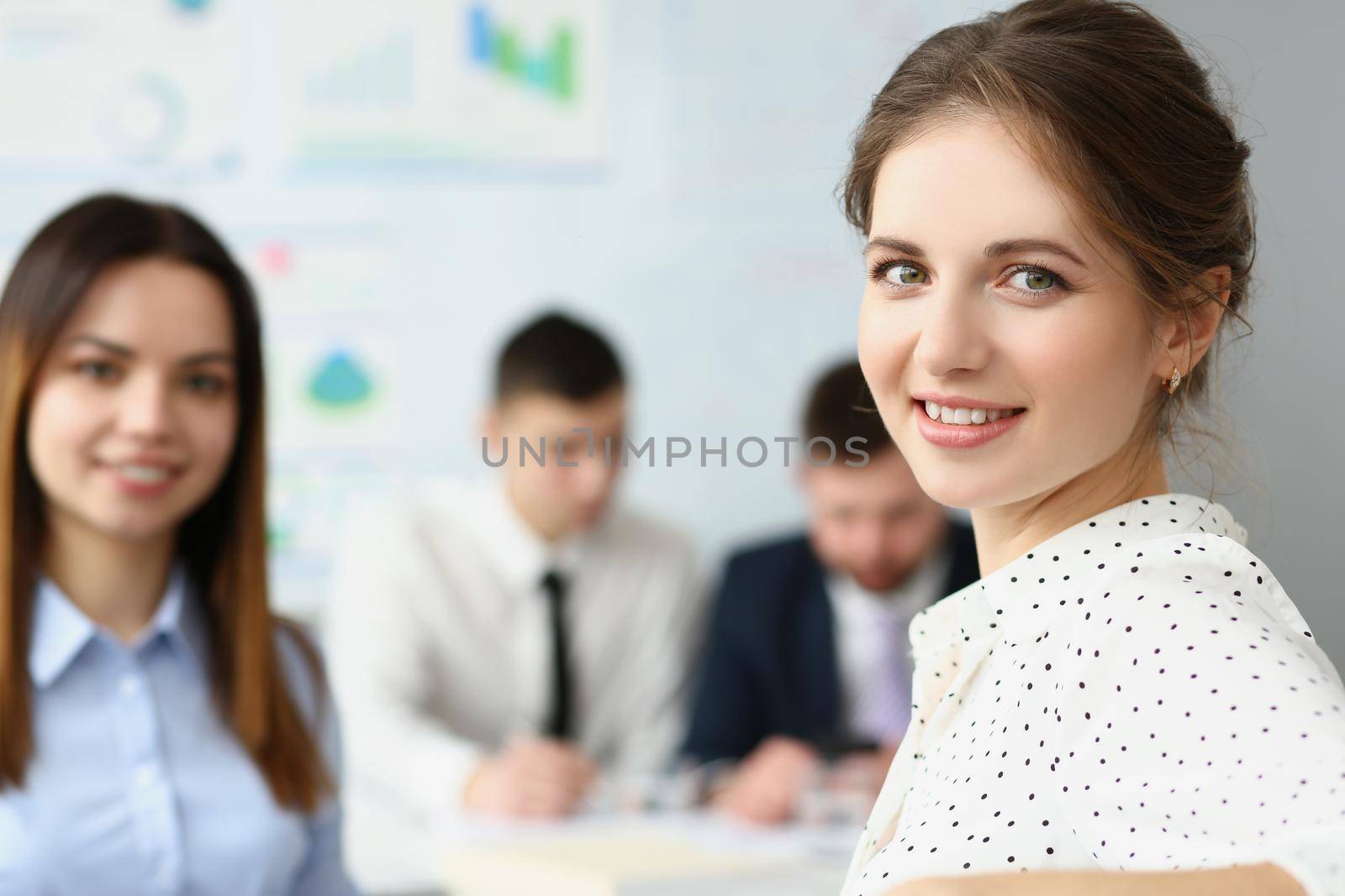 Portrait of smart woman sitting in big modern office with colleagues and smile at camera. Brunette secretary work in friendly collective. Business concept